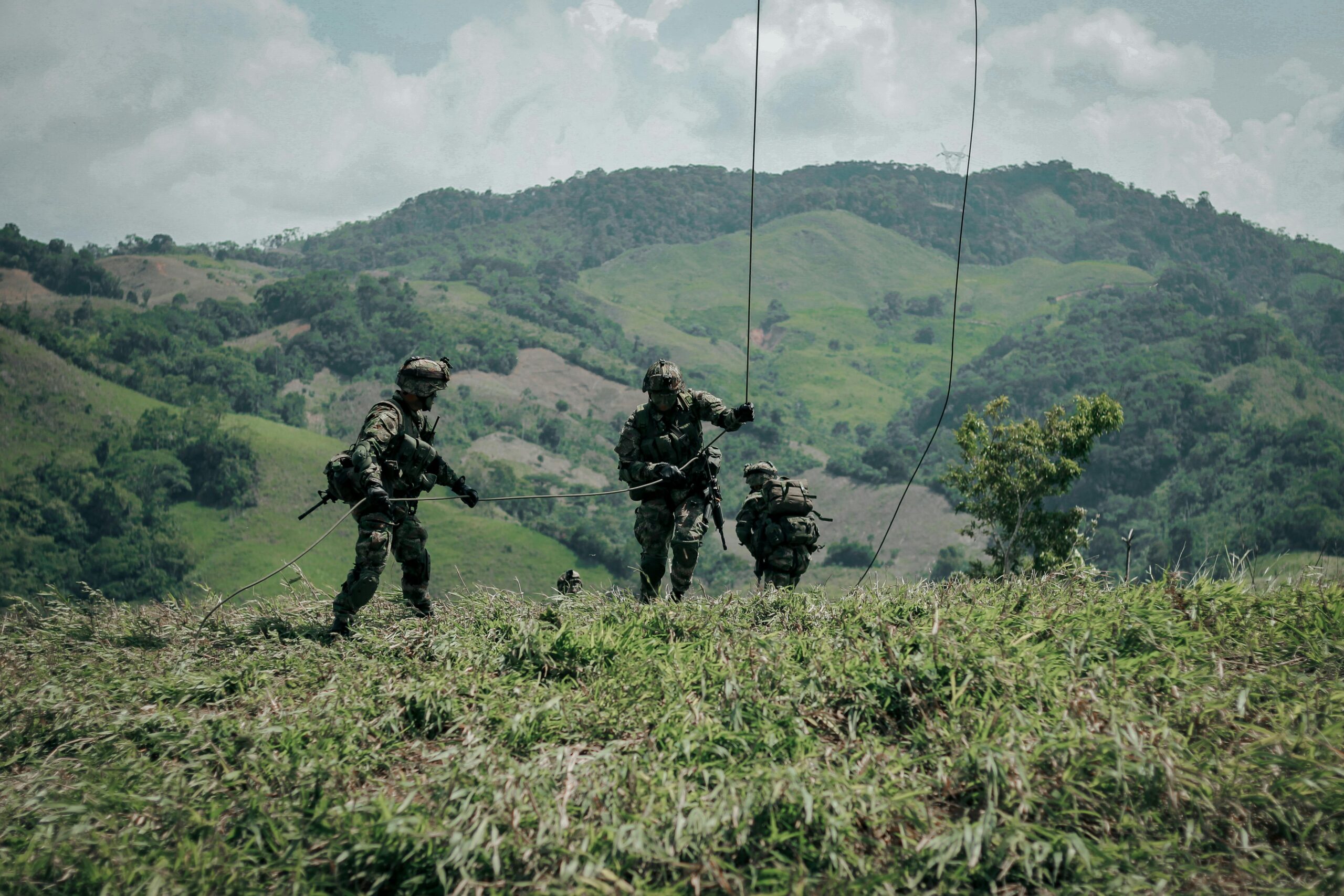 Three soldiers in military gear navigate grassy hills in Colombia during a tactical exercise.
